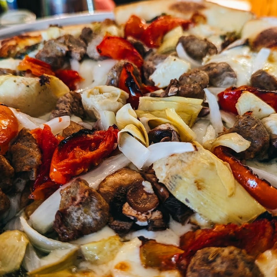 Close-up of a pizza with sausage, roasted red peppers, onions, mushrooms, and artichoke hearts.