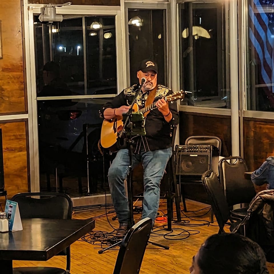 A man plays an acoustic guitar on a small stage in a restaurant with patrons seated at tables.